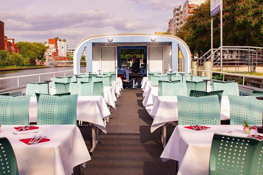 Un espace repas sur un bateau, avec des tables et des chaises, une personne à l’arrière-plan et des bâtiments le long de la rivière. 5