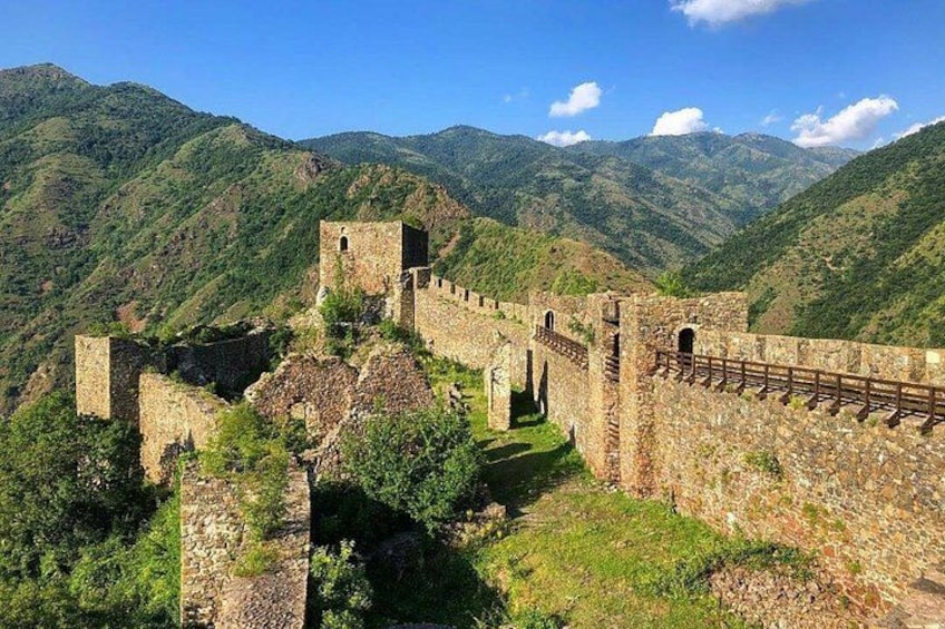 A historical stone wall with towers, set against a mountainous backdrop. 1