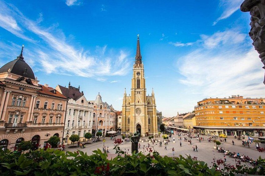 A historic square with a prominent church tower, surrounding classical architecture, and a clear blue sky. 1
