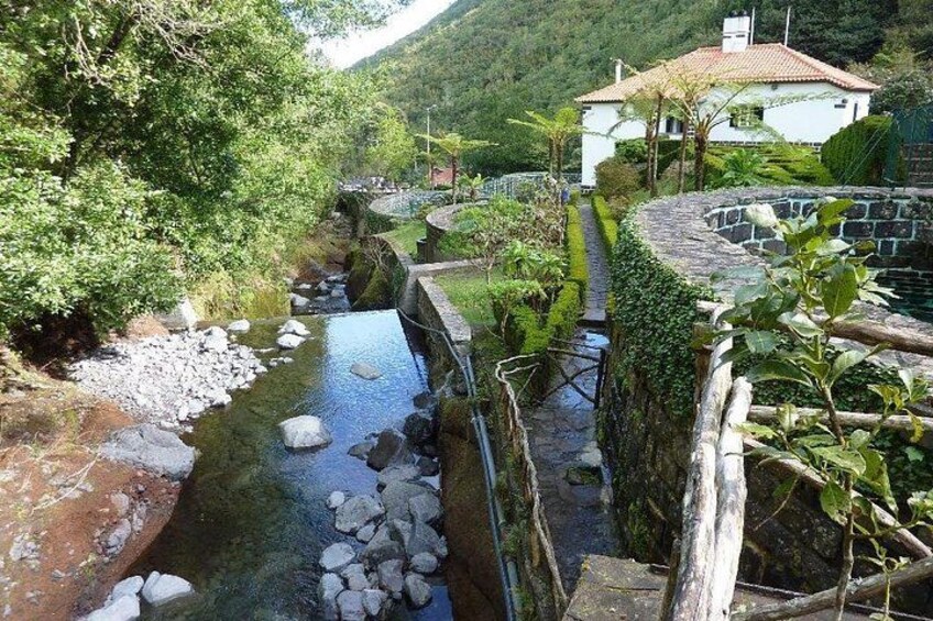 Eine Stebrücke über einen Bach, umgeben von üppiger Vegetation und einem Haus im Hintergrund. 3