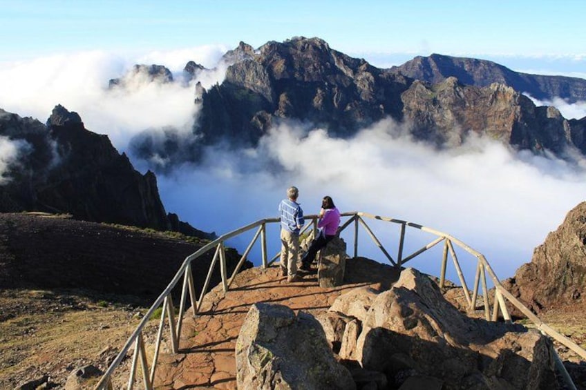 A person sitting on a rocky outcrop with a metal railing, overlooking a mountainous landscape with clouds below. 1