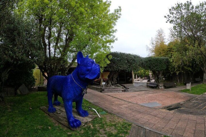 Een blauw hondenbeeld in een tuin met bomen en een pad van bakstenen. 2