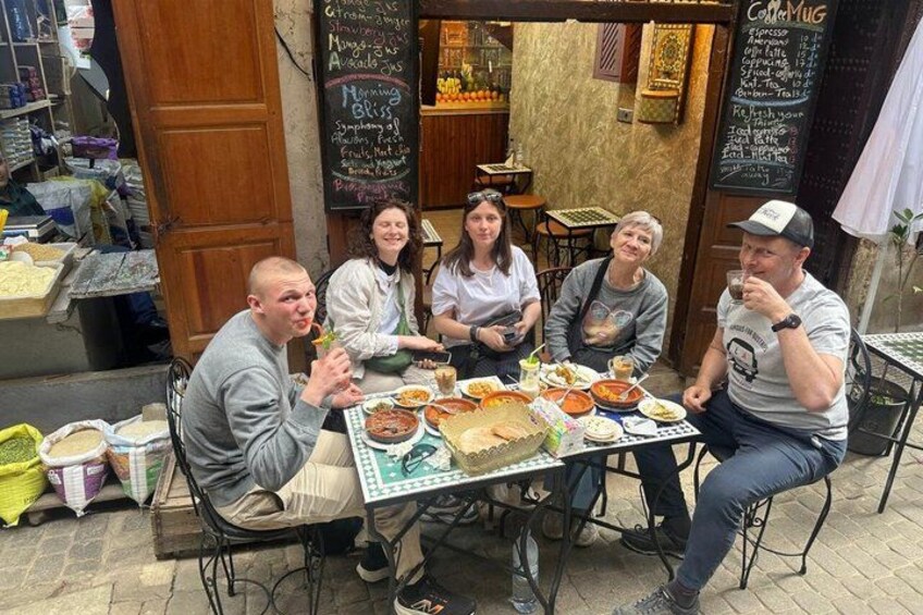 En gruppe mennesker spiser på en udendørs café med flere retter på bordet. 4