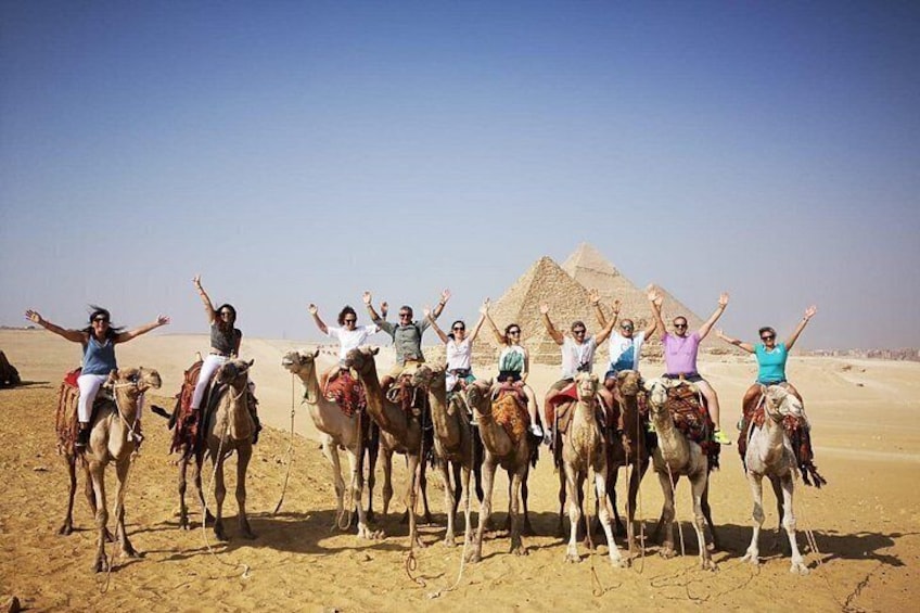 A group of people on camels in a desert with pyramids in the background. 1