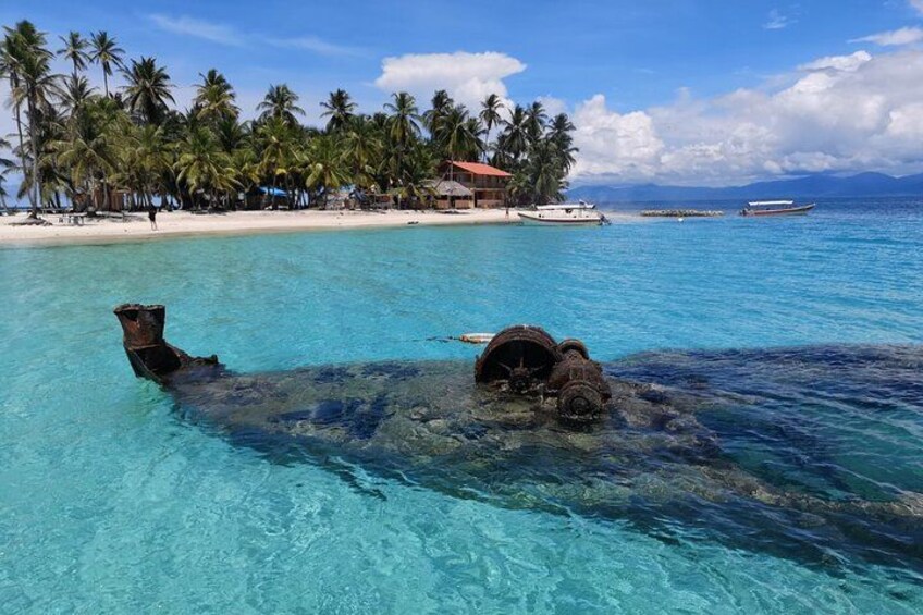 A sunken shipwreck in clear turquoise waters, palm trees, and a beach with buildings in the background. 5