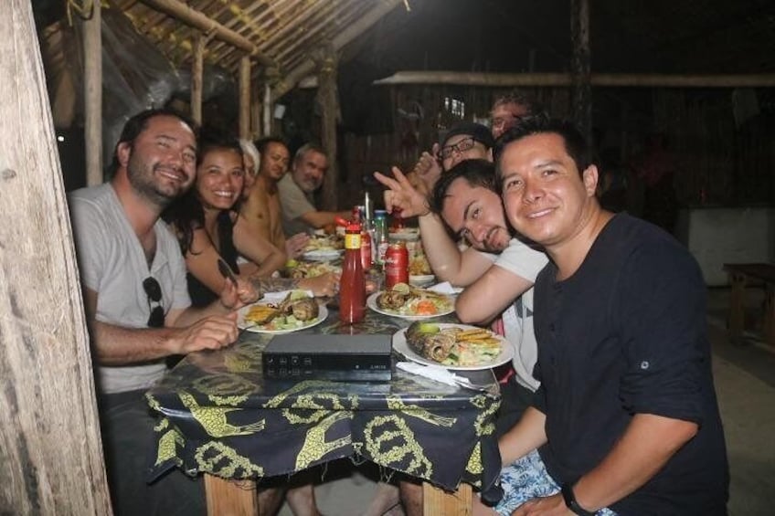 Un groupe de personnes est assis à une table en bois à l’extérieur, une nappe recouvre la table, et on y voit des assiettes remplies de nourriture et des condiments. 1