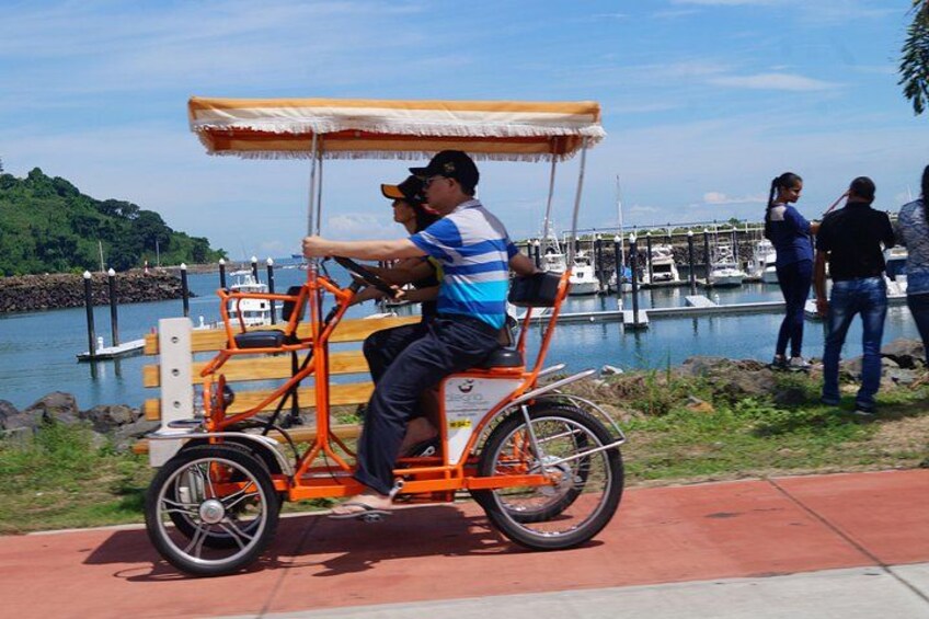 Un homme fait du vélo sur une bicyclette orange munie d’un auvent, près d’une étendue d’eau où se trouvent des bateaux et un quai. 4