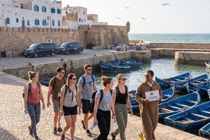 Un groupe de personnes se promène le long d’une promenade en bord de mer, avec des bateaux amarrés à proximité et un bâtiment historique en arrière-plan. 3