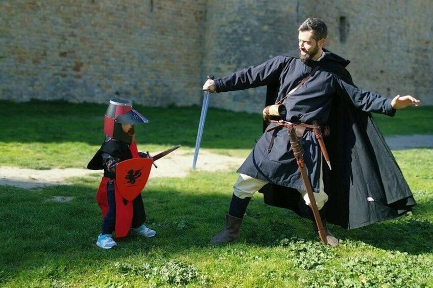Una persona vestida con traje medieval, con espada y escudo, de pie en un área cubierta de hierba, con un muro de piedra al fondo. 2