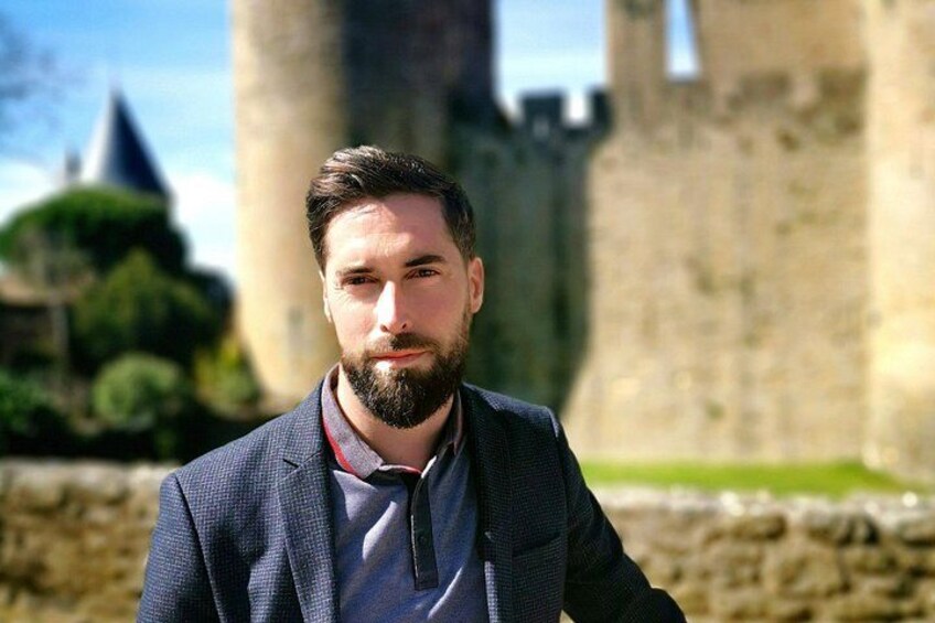 A man in a suit standing in front of a historic stone wall with a castle in the background. 2