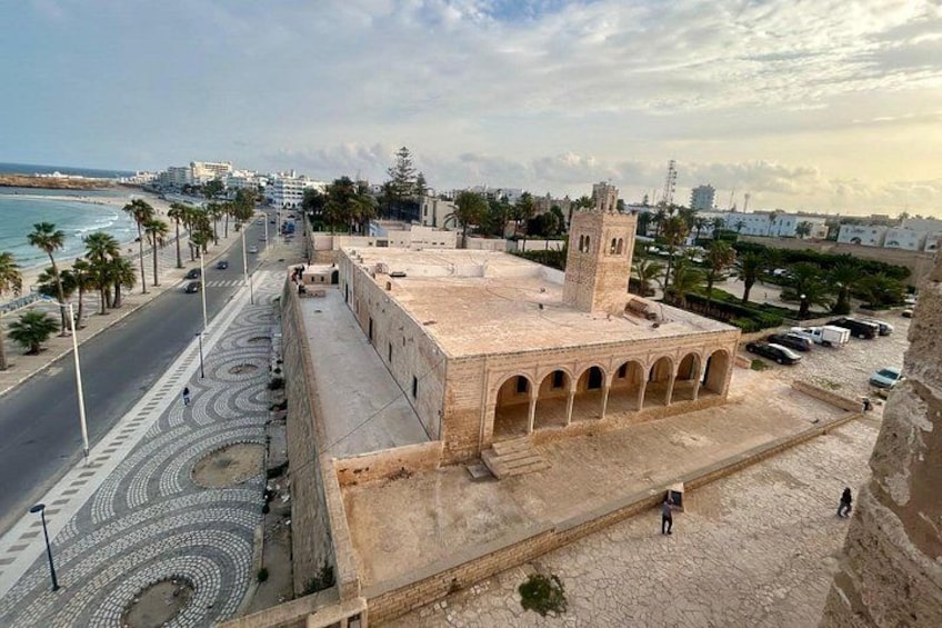 Un paisaje urbano costero con un edificio histórico destacado que presenta arcos y una torre, junto a una carretera moderna con palmeras y una playa al fondo. 1