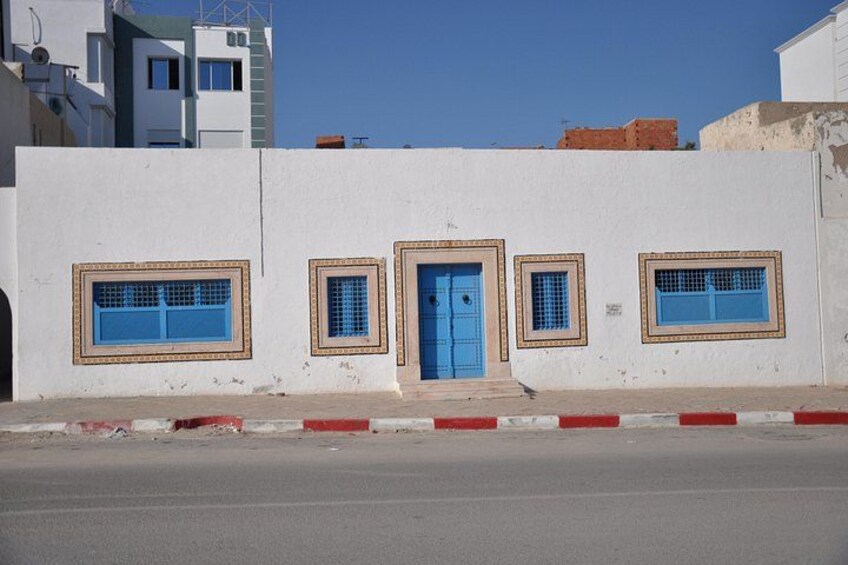 Un edificio blanco con puertas y ventanas azules, una acera roja y blanca, y un cielo azul despejado. 4