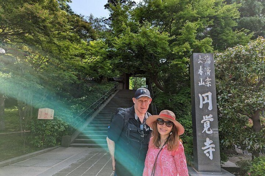 Zwei Personen stehen vor einem Schild mit japanischen Schriftzeichen, umgeben von viel Grün und einer Treppe, die zu einem überdachten Bereich führt. 2