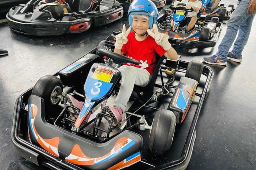 A child in a go-kart with number 3, wearing a blue helmet and a red shirt, at a go-kart track. 3