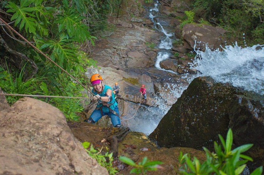 A person rappelling down a rocky cliff with a rope, surrounded by lush greenery and a flowing stream. 3