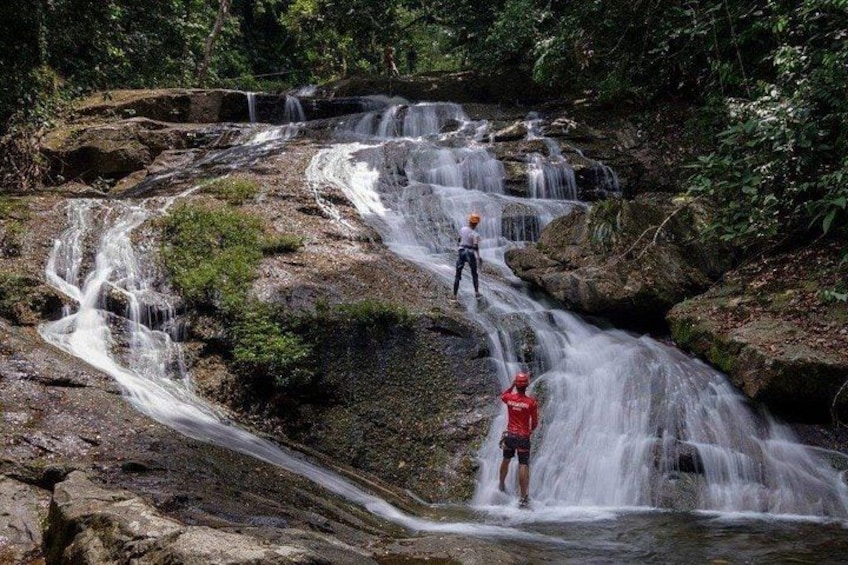 A waterfall with multiple streams, a person in a red shirt standing on a rock, and another person in a white shirt and helmet observing. 1