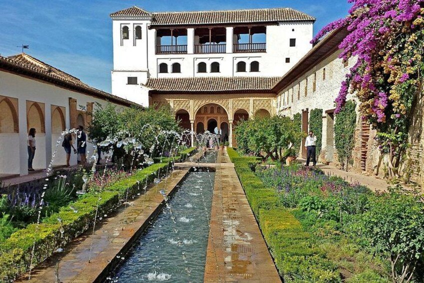 Un patio con un canal de agua, un edificio con balcones y plantas florecientes. 4