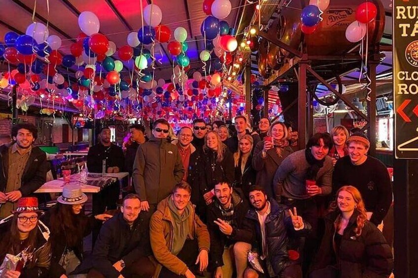 A group of people in a festive indoor setting with balloons and a bar area. 2