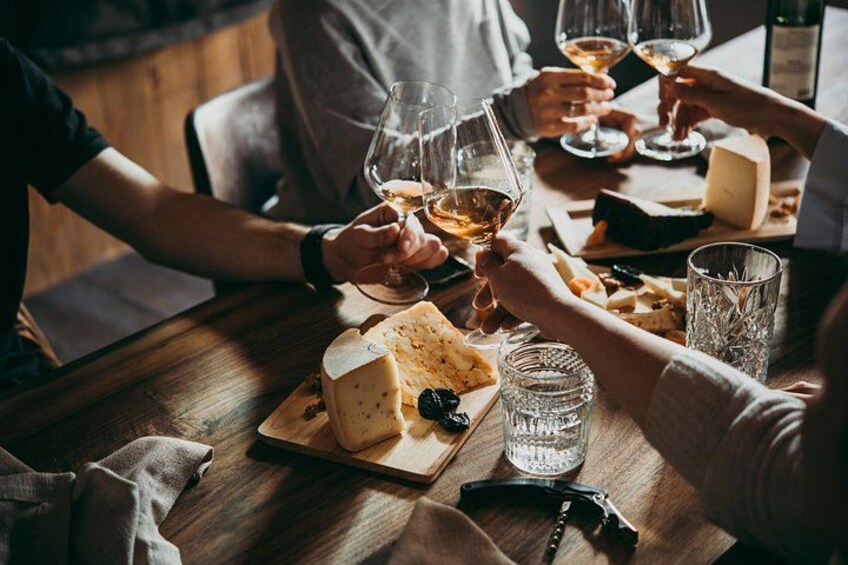 A group of people toasting with wine glasses at a wooden table with cheese and bread. 1