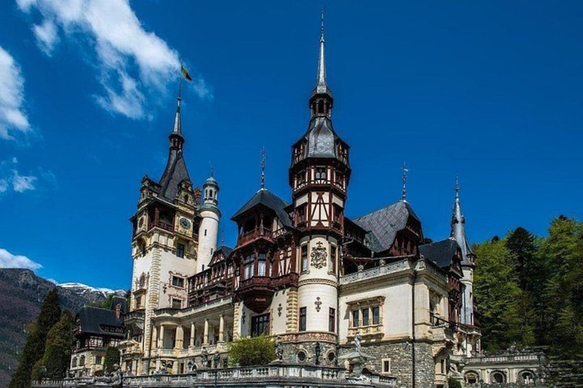 A historic castle with multiple towers and a prominent clock tower, surrounded by a mountainous landscape. 3