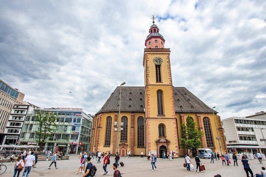 A historic church with a clock tower, surrounded by modern buildings and a busy square. 2