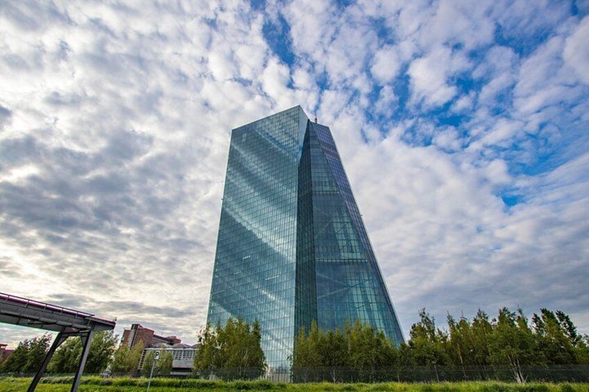 A tall glass skyscraper with a reflective surface, surrounded by trees and a partly cloudy sky. 4