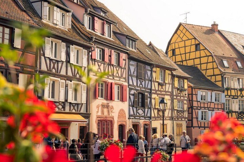 A European town with colorful half-timbered houses, a central building with a red facade, and people walking in the foreground. 1