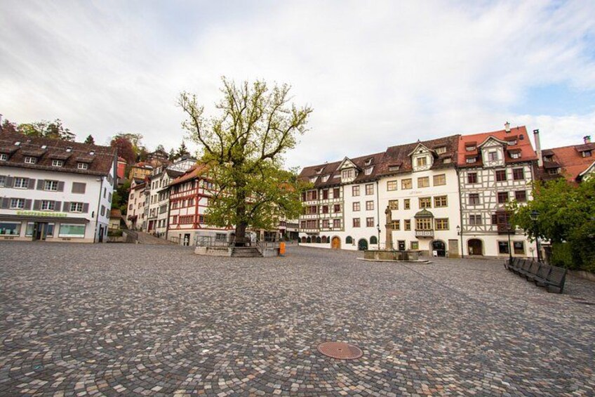Ein Kopfsteinpflasterplatz mit historischen Gebäuden, einem Baum und einer Bank. 2