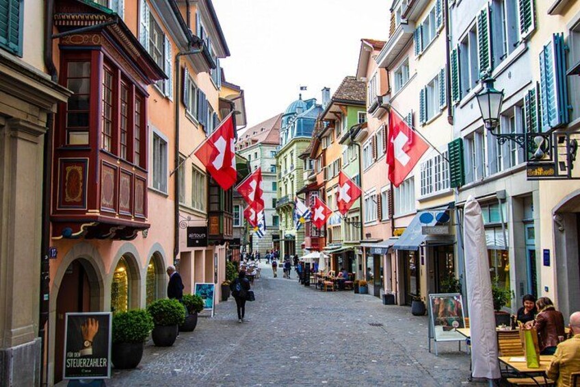 A cobblestone street lined with colorful buildings, Swiss flags, and outdoor seating areas. 2