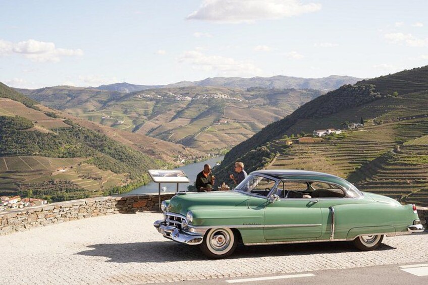 A green vintage car parked on a cobblestone road with a scenic view of rolling hills and a body of water in the background. 1