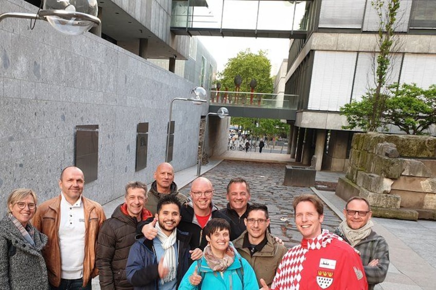 A group of people standing in an urban outdoor setting with modern architecture, including a stone wall and a glass bridge. 3