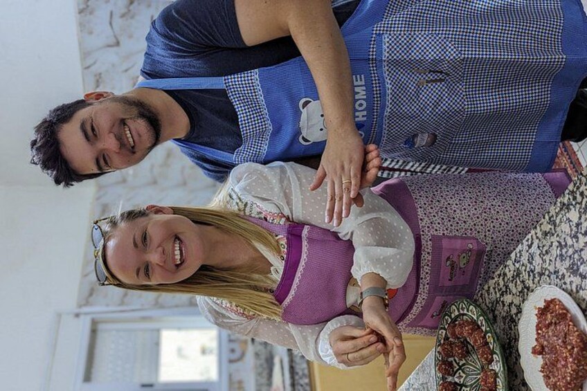 Two people wearing aprons, one with a blue apron and the other with a purple apron, standing in a kitchen with a table full of food. 5