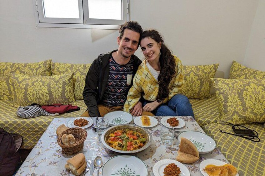 A couple sitting on a patterned couch with a table in front of them, filled with plates of food and a basket. 4