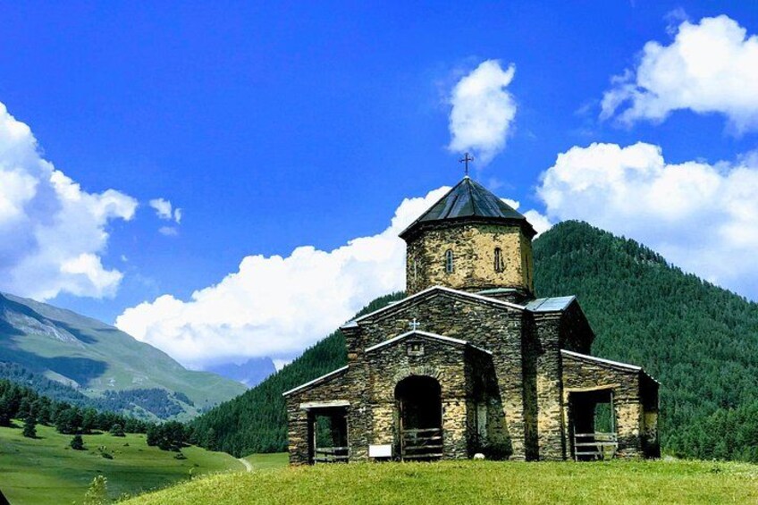 A stone church with a bell tower, surrounded by a green landscape and mountains. 2