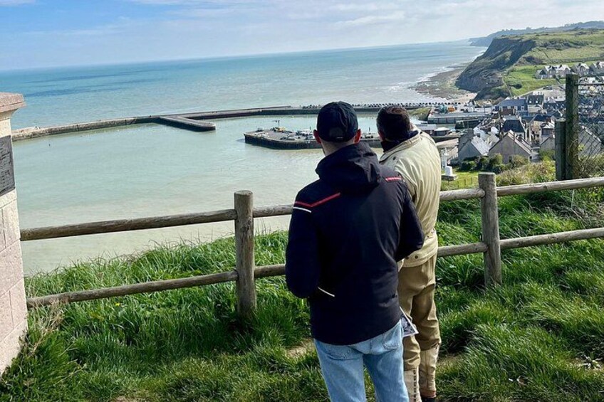 Deux personnes se tiennent sur une colline herbeuse, d’où elles dominent une ville côtière et ont une vue dégagée sur la mer et une digue. 2