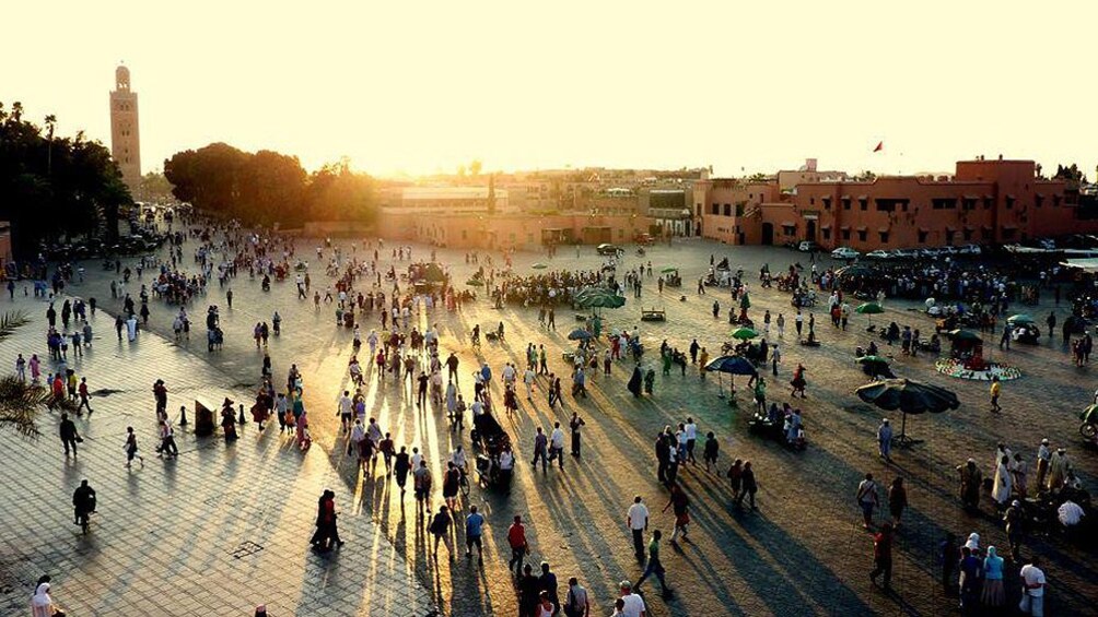 A bustling square with people walking and sitting, surrounded by buildings and a prominent tower. 1