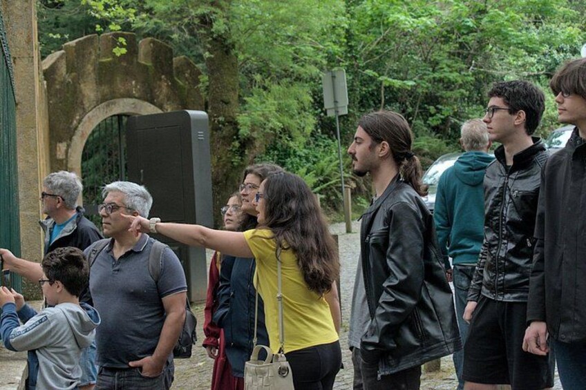 A group of people standing outdoors, some pointing at something, with greenery and a stone structure in the background. 2