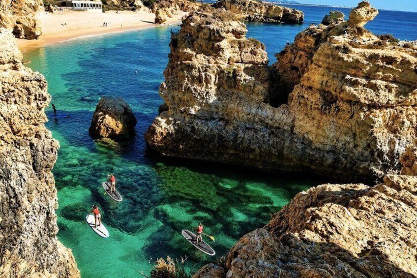 A rocky coastal area with clear turquoise water, a sandy beach, and people on paddleboards. 3