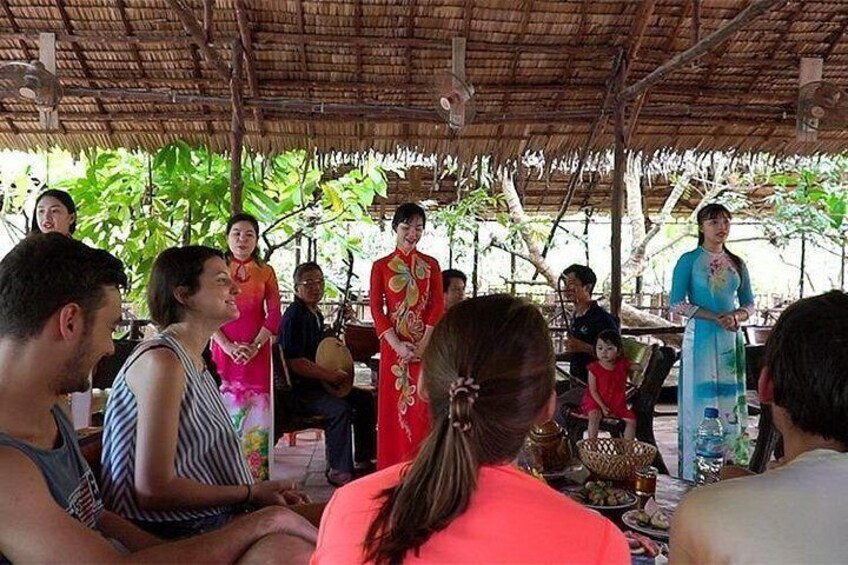 Een groep mensen zit onder een rieten dak, bezig met een culturele activiteit, met een vrouw in traditionele kledij in het midden. 5