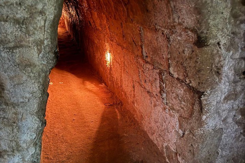 Un túnel estrecho y tenuemente iluminado, con paredes rugosas de color marrón rojizo y una única fuente de luz al final. 3