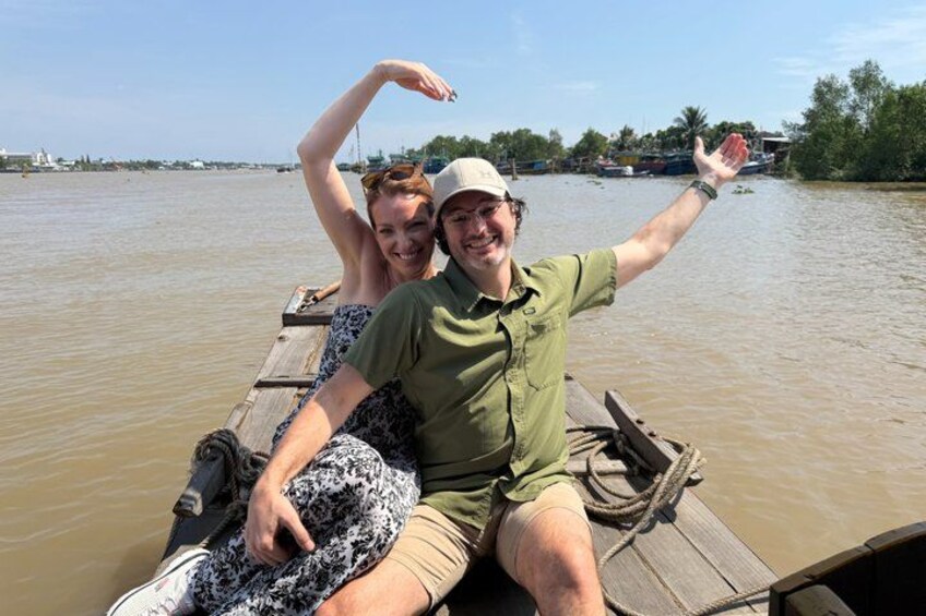Two people on a wooden boat on a river with trees and buildings in the background. 3