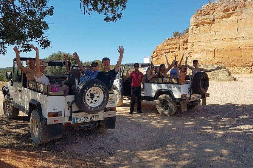 Two off-road vehicles with passengers raising their hands, parked on a dirt road with a rocky cliff in the background. 3