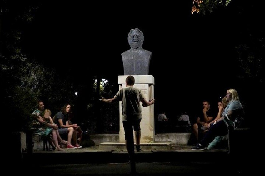 A person standing in front of a bust statue, addressing an audience seated on benches. 4
