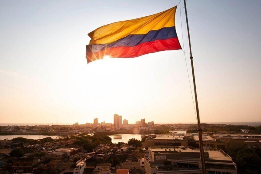 A flag waving in the wind, a city skyline in the background, and a body of water in the foreground. 1