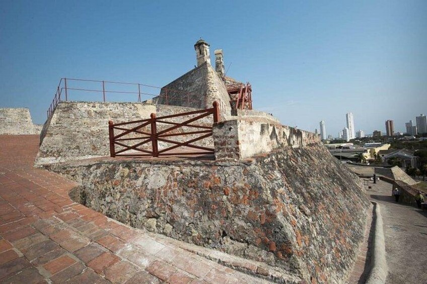 A historical stone fortification with a red gate, a brick pathway, and a cityscape in the background. 5