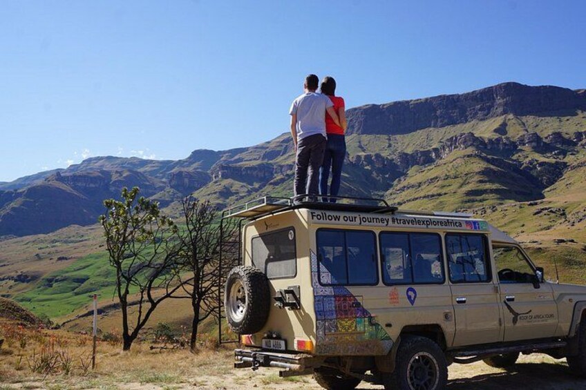 A beige off-road vehicle with a group of people standing on top, a clear sky, and a mountainous landscape. 1