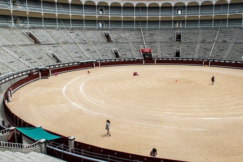 An empty bullring with a sandy floor and tiered seating. 5