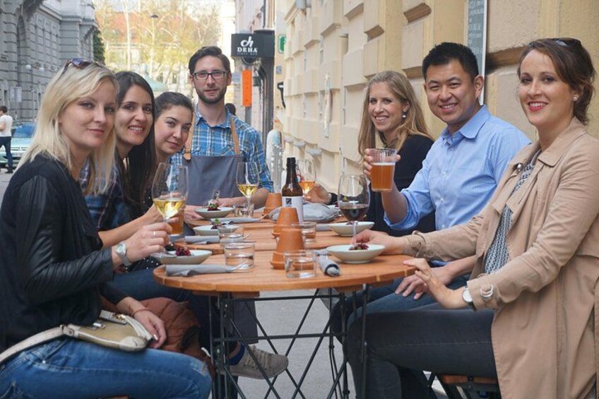 Een groep mensen zit aan een tafel met drankjes en eten, in een openlucht omgeving met gebouwen op de achtergrond. 2