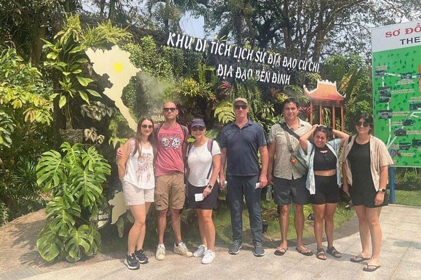 A group of people posing in front of a sign with Vietnamese text, surrounded by lush greenery and a traditional pavilion. 4