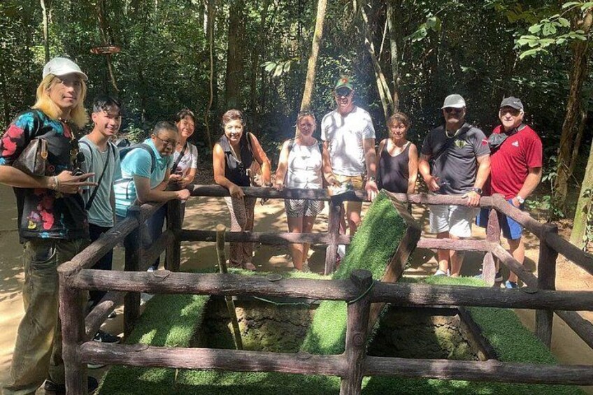 A group of people standing behind a wooden fence with a green artificial grass barrier, in a forested area. 2